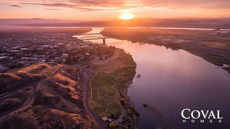An aerial view of a wide river at sunrise or sunset under a vibrant orange and purple sky. On the left bank, a city and a large green park with winding paths sit adjacent to rugged hills. A large bridge spans the river in the distance, with the sun glowing brightly on the horizon. The logo "COVAL HOMES" is displayed in the bottom right corner.