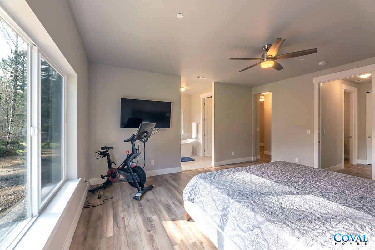 Spacious bedroom with king bed, wall-mounted TV, exercise bike, and large windows overlooking trees