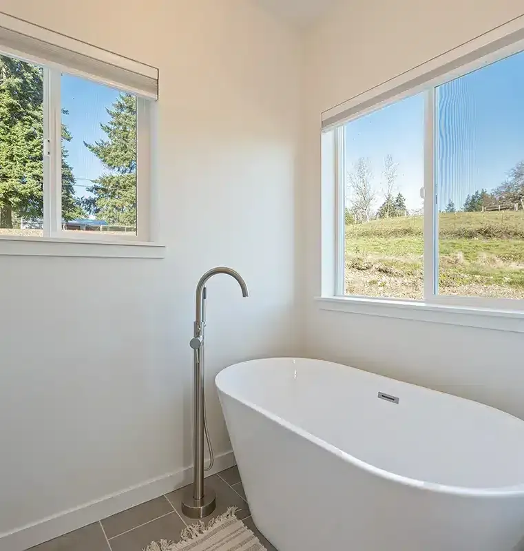 Bright bathroom with a freestanding white tub, floor-mounted faucet, and two windows overlooking greenery