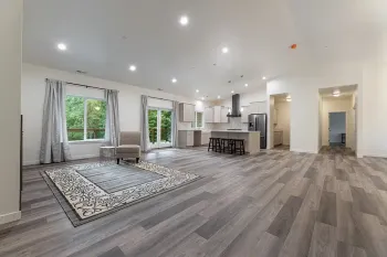 Bright open living room with gray floors, white walls, large windows, rug, and kitchen island.