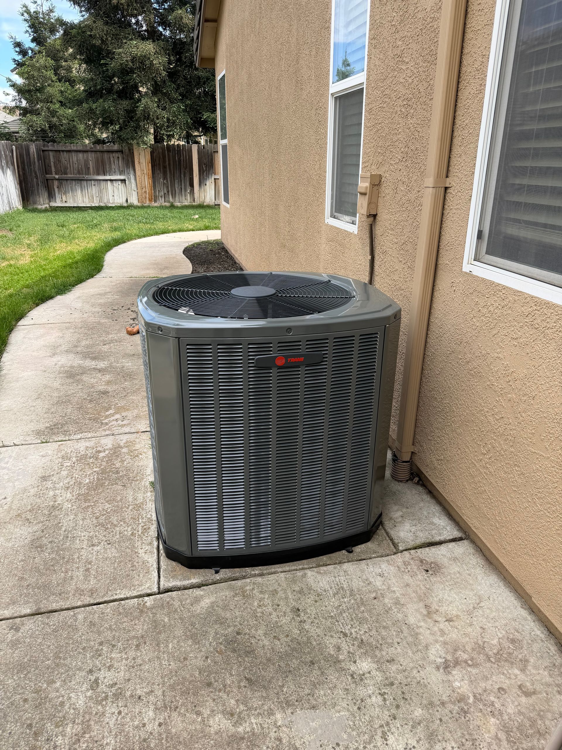 A light gray outdoor residential air conditioning unit sits on a concrete pad against a beige exterior wall.