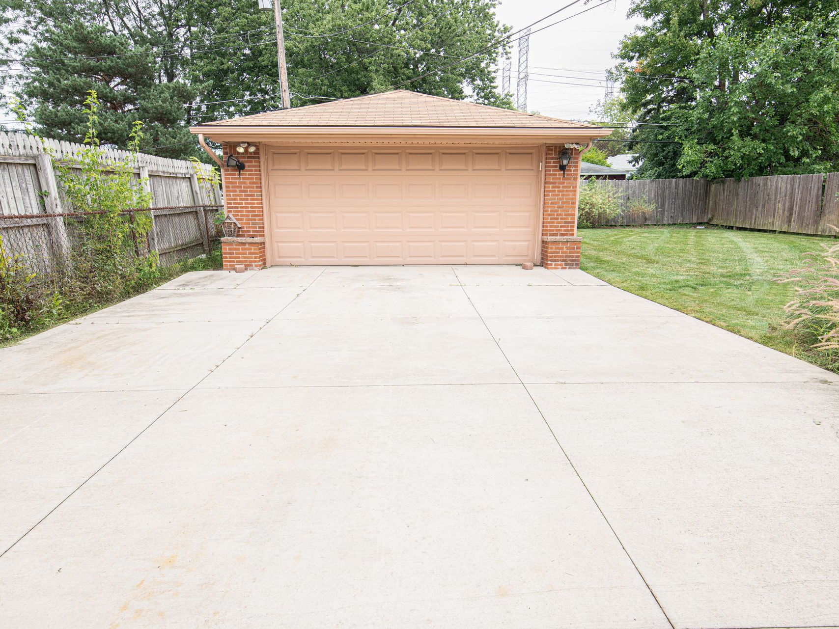 Single-car garage with tan door and wide concrete driveway, bordered by brick walls and grass.