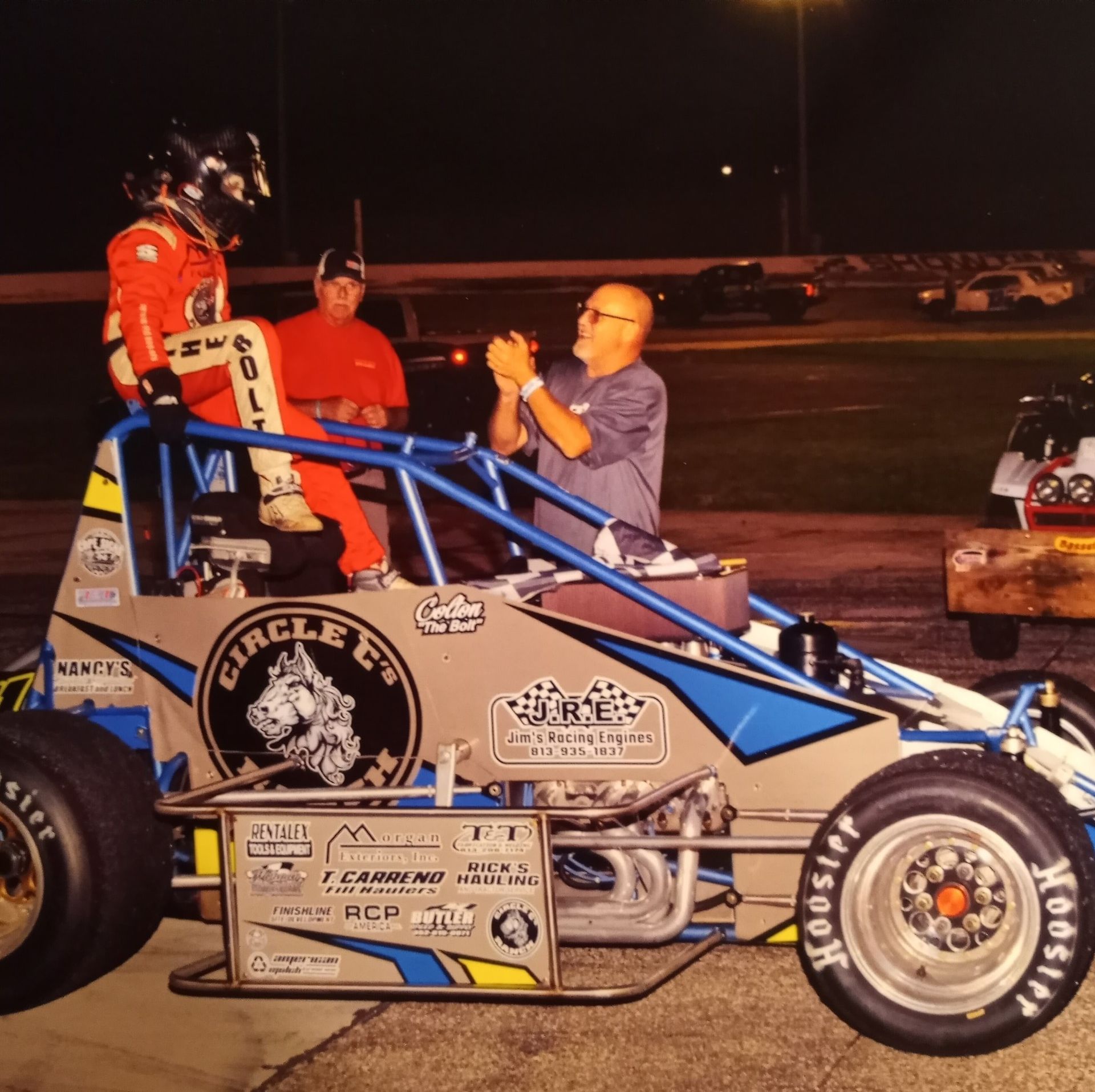 Race car driver celebrates win on his car with crew, night setting.