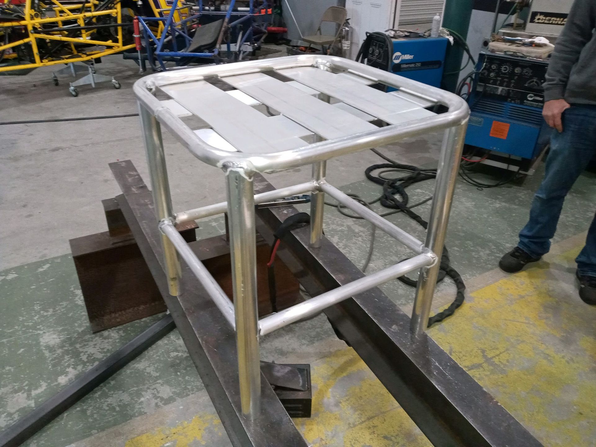 Aluminum welded cart frame, resting on a steel beam in a workshop, with a person standing nearby.