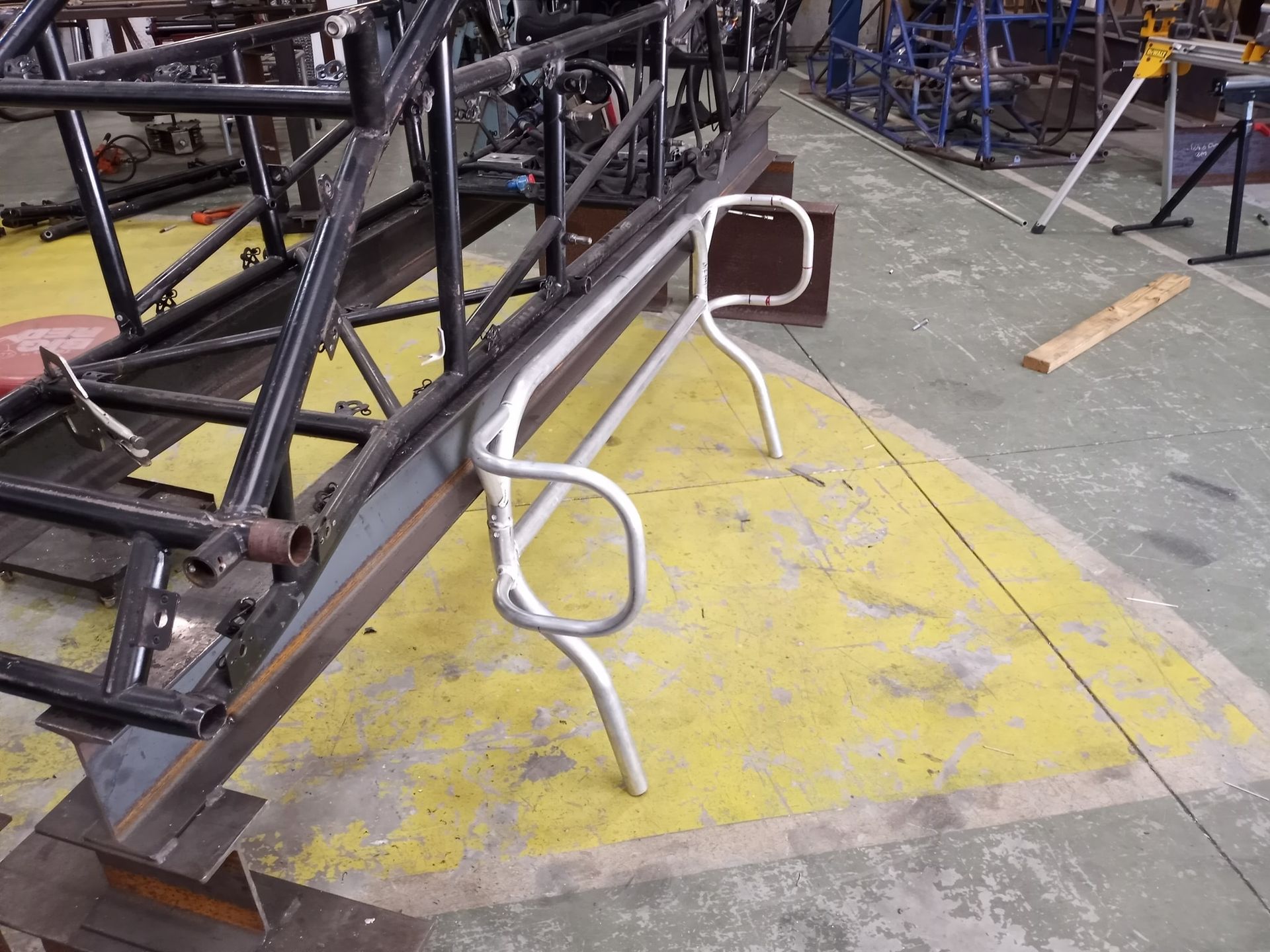 A steel chassis with a curved metal bar welded to it, on a yellow painted floor in a workshop.