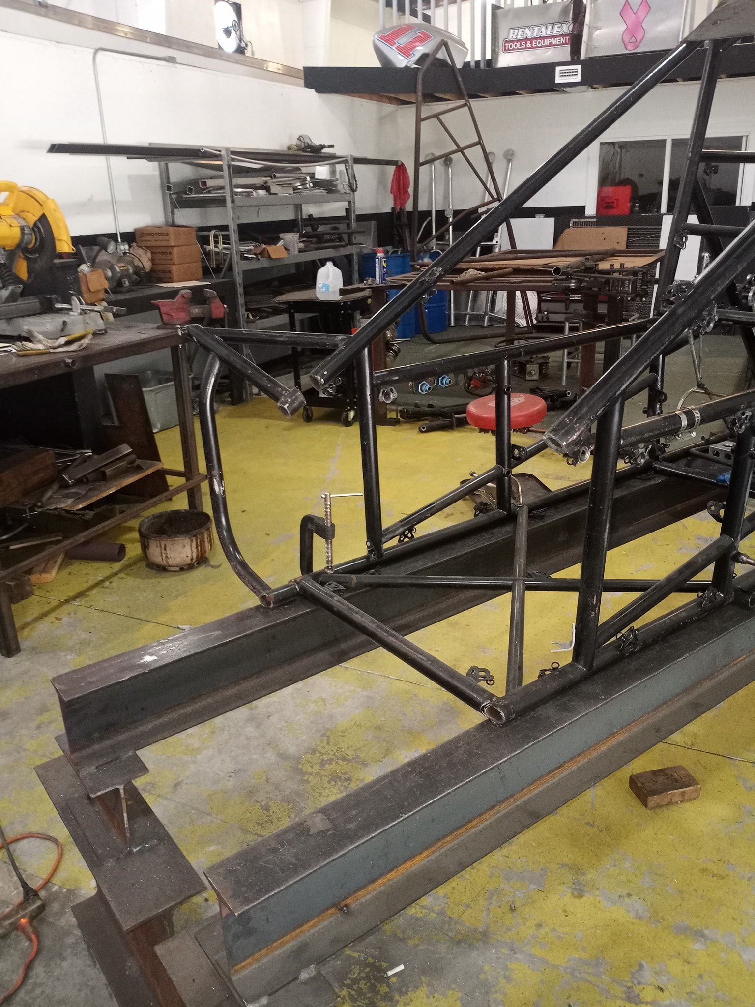 Metal frame of a vehicle being constructed in a workshop; welding tools, materials visible in the background.