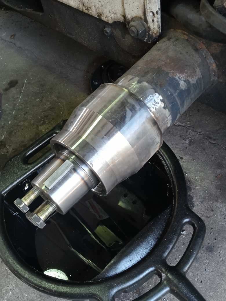 Metal component with bolts, placed in a black oil drain pan; dark setting with hints of rust.