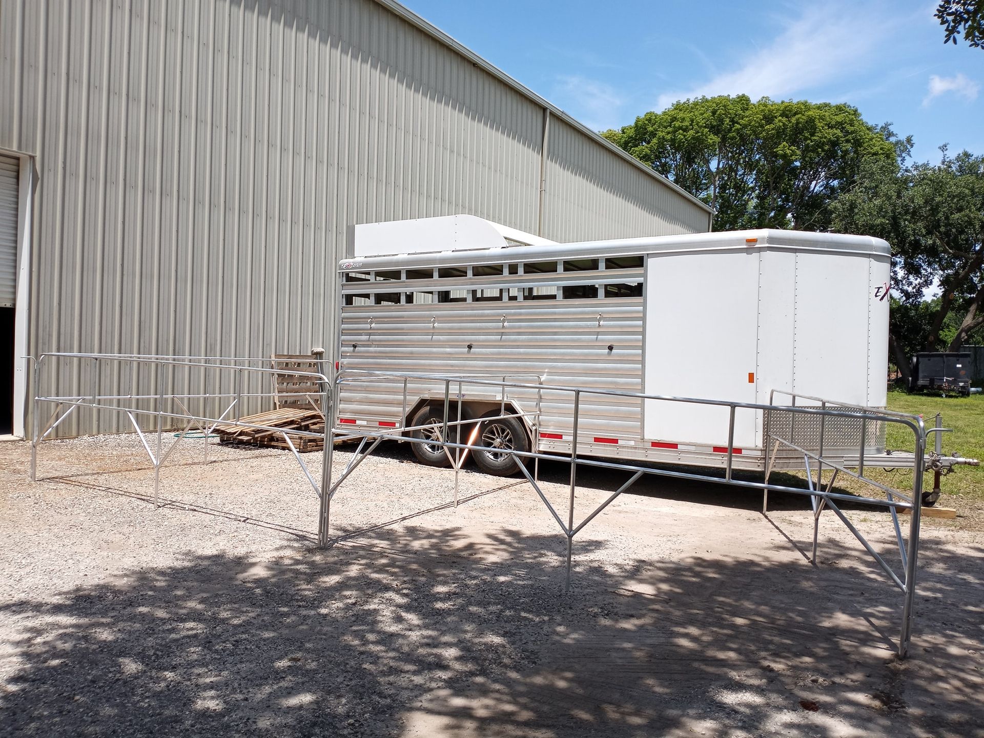 Horse trailer parked near a corrugated metal building, surrounded by metal fencing, gravel ground.