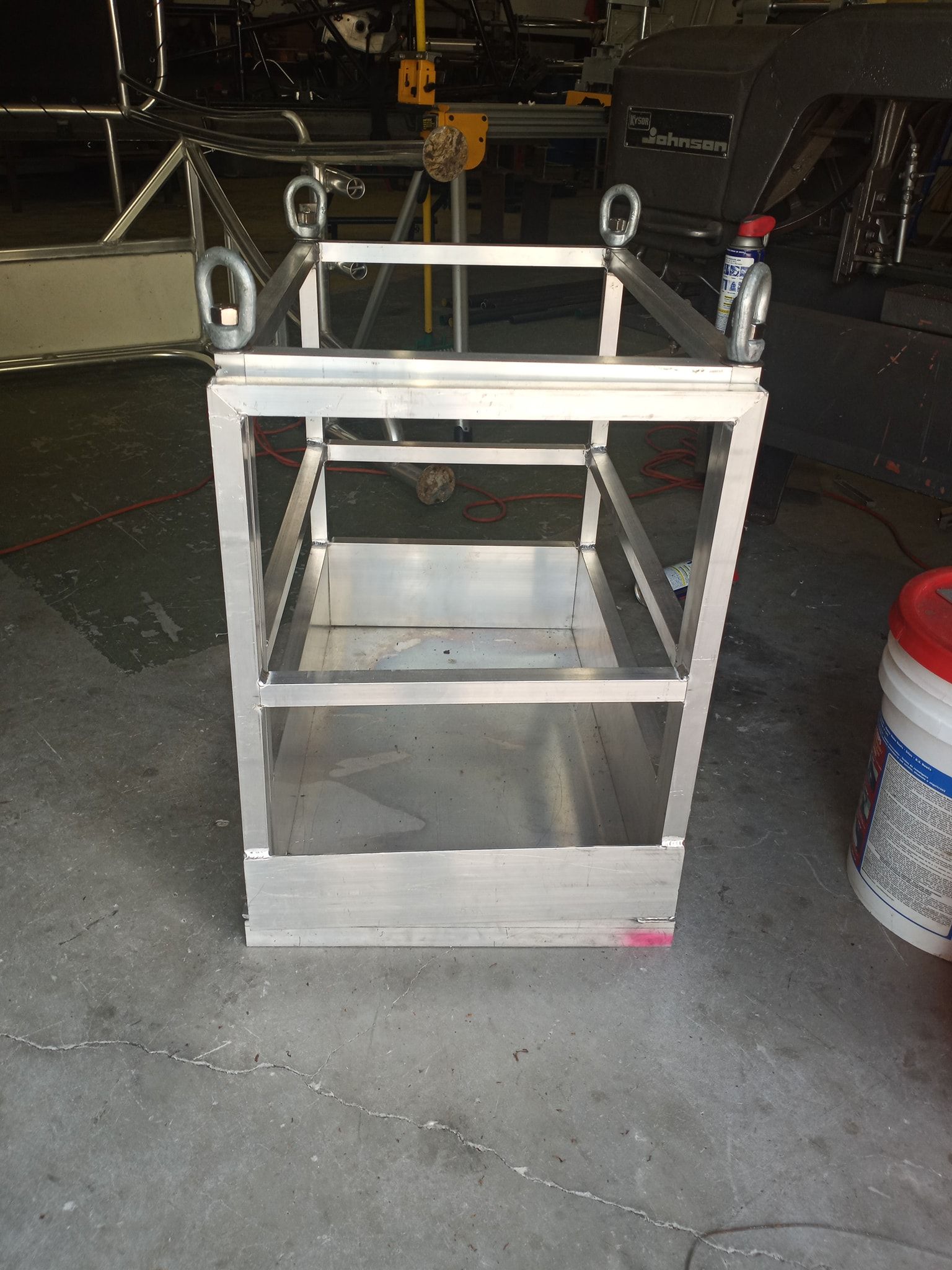 Metal lifting basket with open top, solid bottom, and eye bolts for attachment.