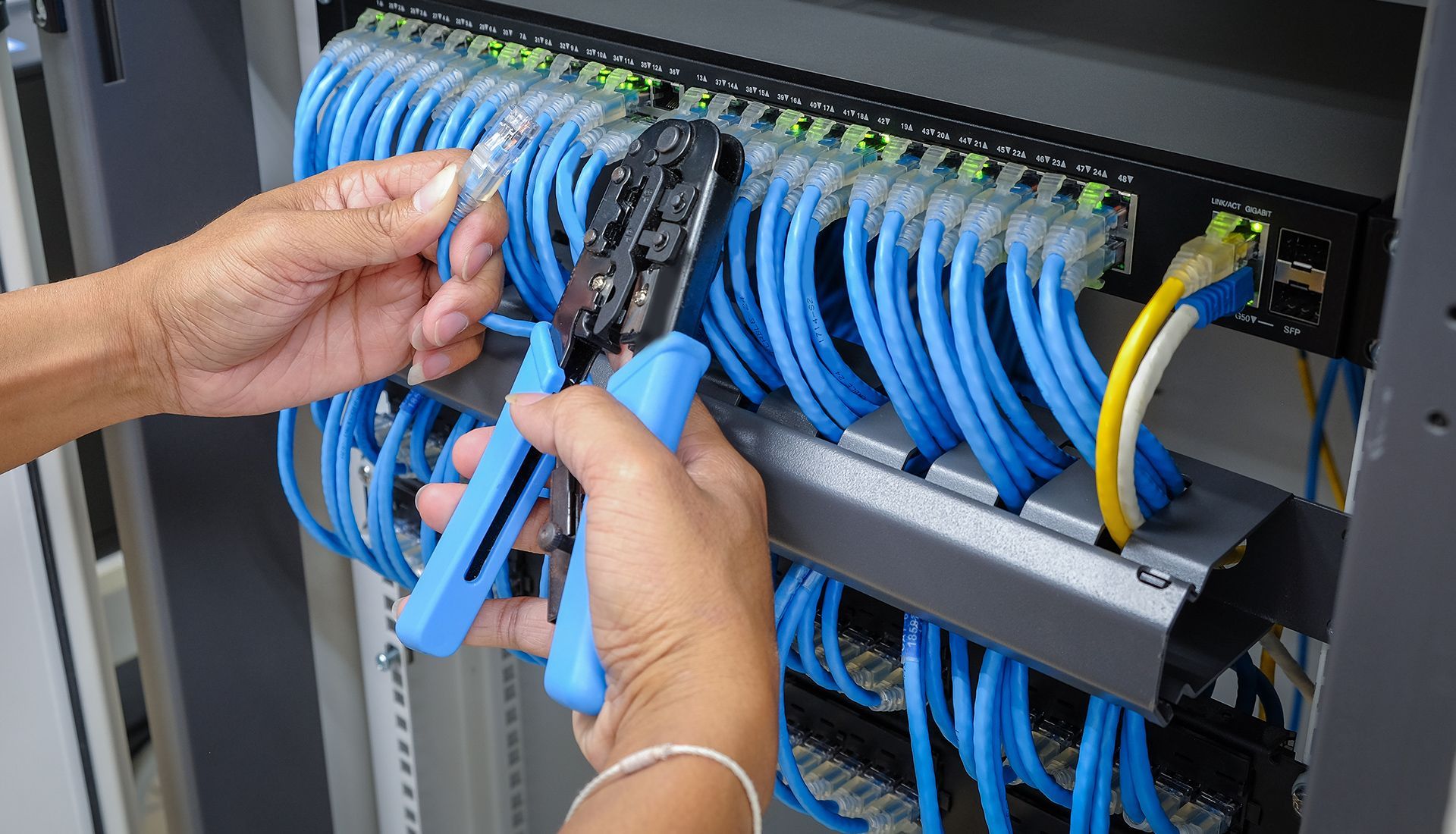 Hands using crimping tool to connect blue Ethernet cables to a network switch in a server rack.
