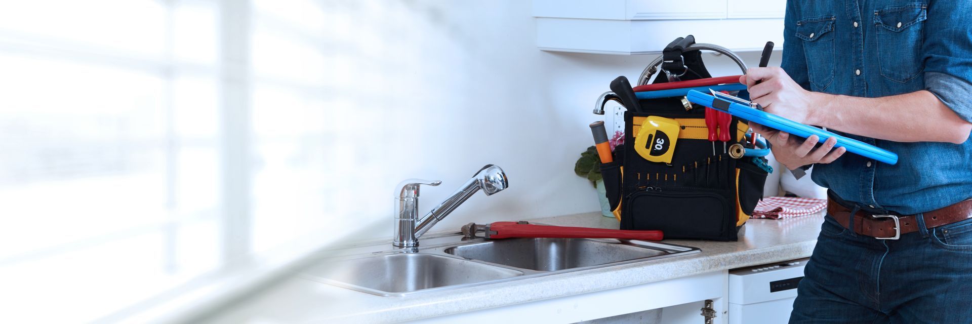 Plumber reviewing a clipboard beside a kitchen sink with tools and a tool bag on the counter. Plumber reviewing a clipboard beside a kitchen sink with tools and a tool bag on the counter.