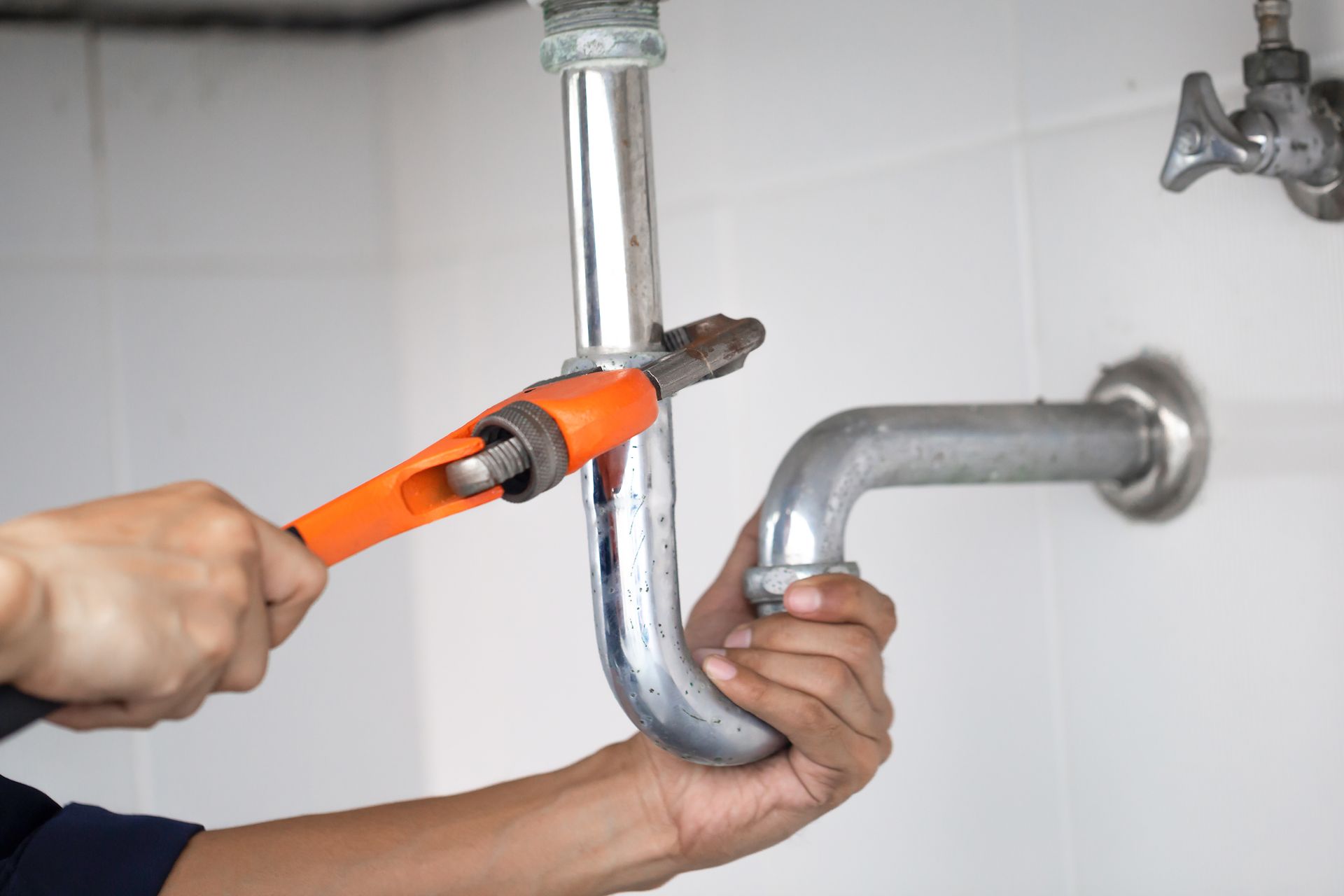 A person uses an orange pipe wrench to tighten a metal P-trap pipe assembly under a sink.