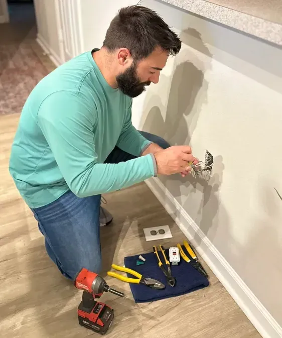 Man in blue shirt kneels, wiring an outlet with tools on a cloth, next to a white wall and floor.