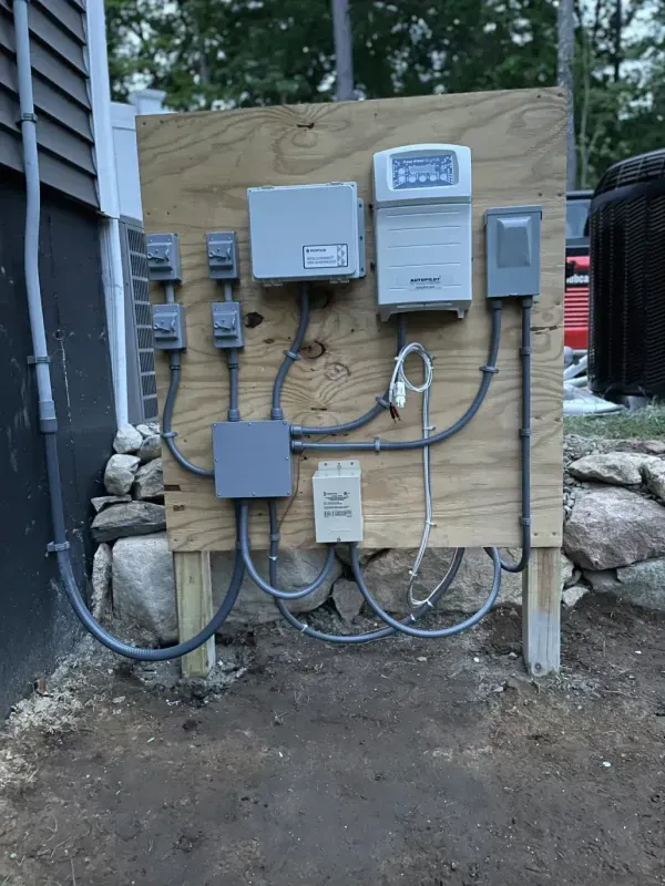 Outdoor electrical panel with various gray boxes, wiring, and control panel mounted on a wooden board.