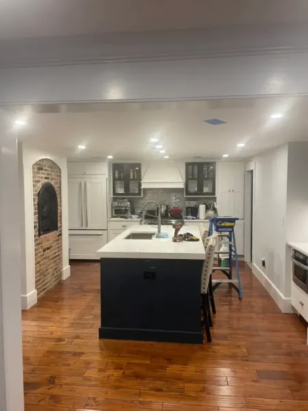A modern kitchen with a blue island, white cabinets, brick accent wall, and wood floors.