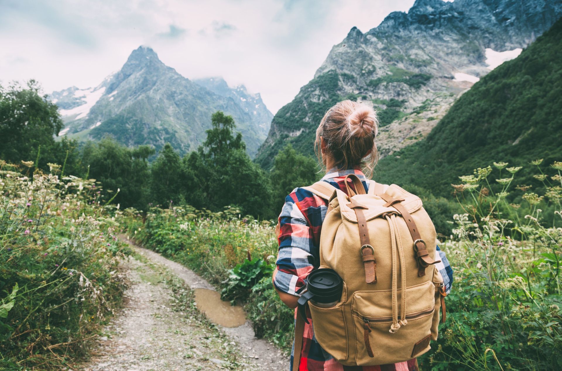 Woman with backpack hikes on a trail, viewing snow-capped mountains.