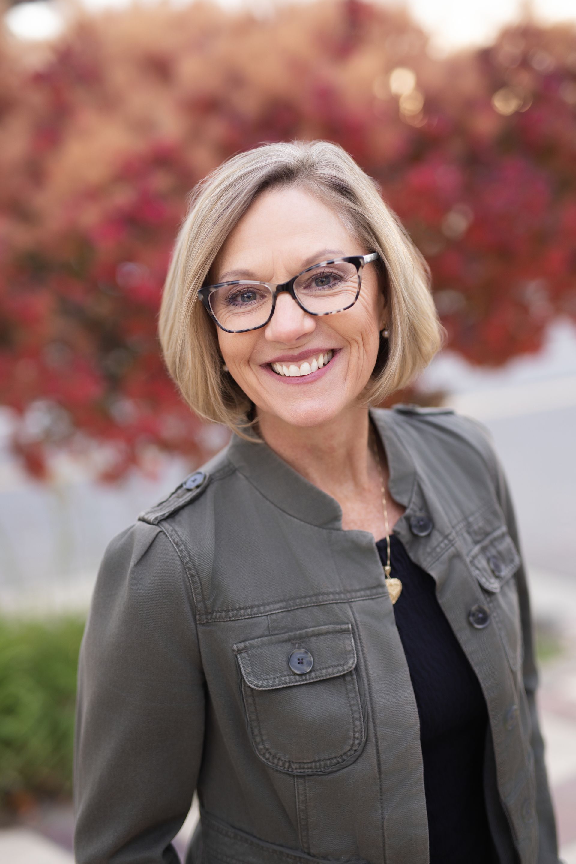 Joy Mitchell wearing glasses smiles at the camera, wearing a green jacket, in front of fall foliage.