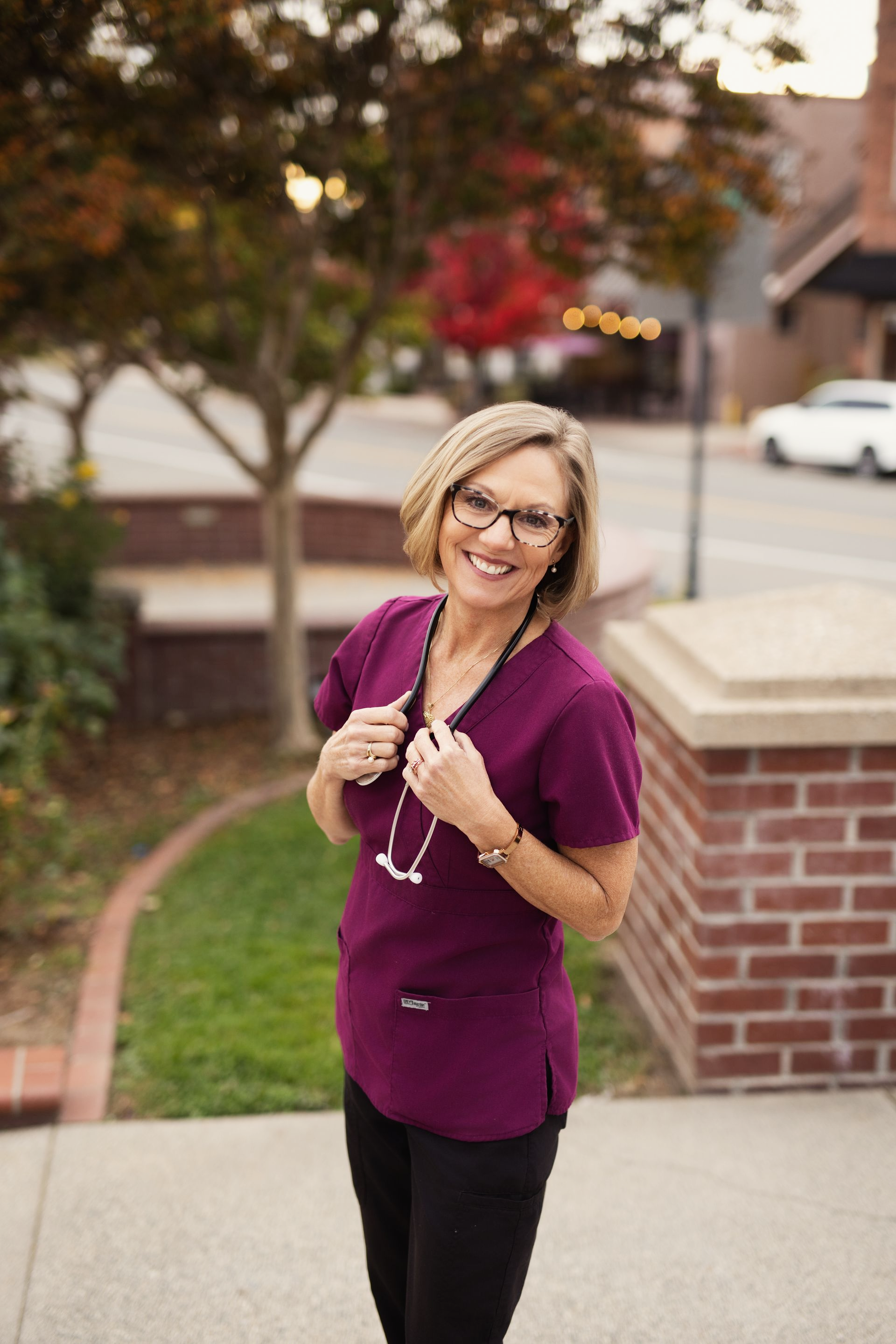 Joy Mitchell in purple scrubs and glasses smiles outdoors.