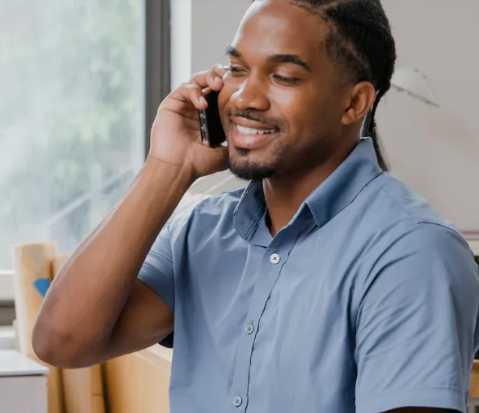 Homem ao telefone, sorrindo. Vestindo camisa azul de botões. Perto da janela, dentro de casa.