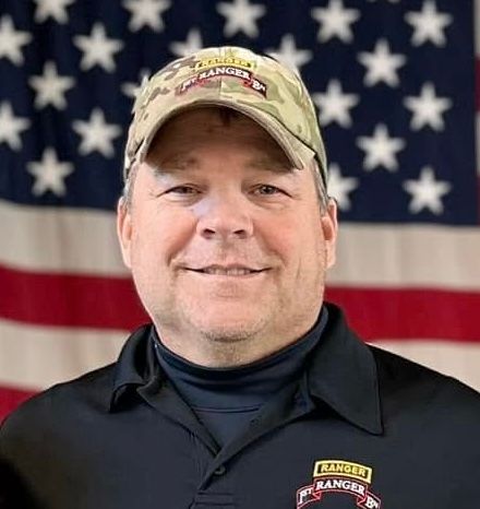 A man wearing a hat and a black shirt is standing in front of an american flag.
