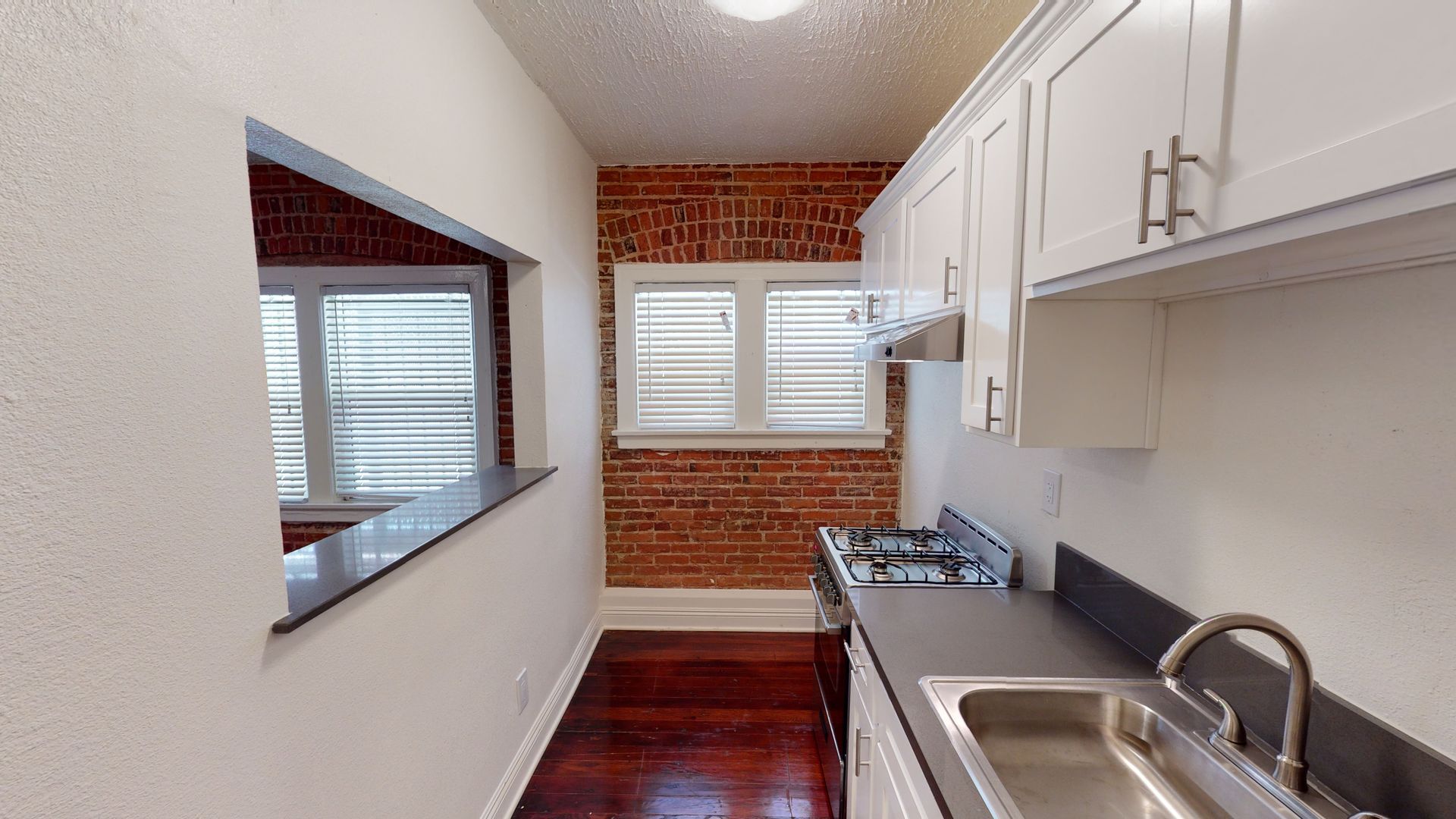 Narrow kitchen with white cabinets, dark countertops, brick wall, and window.