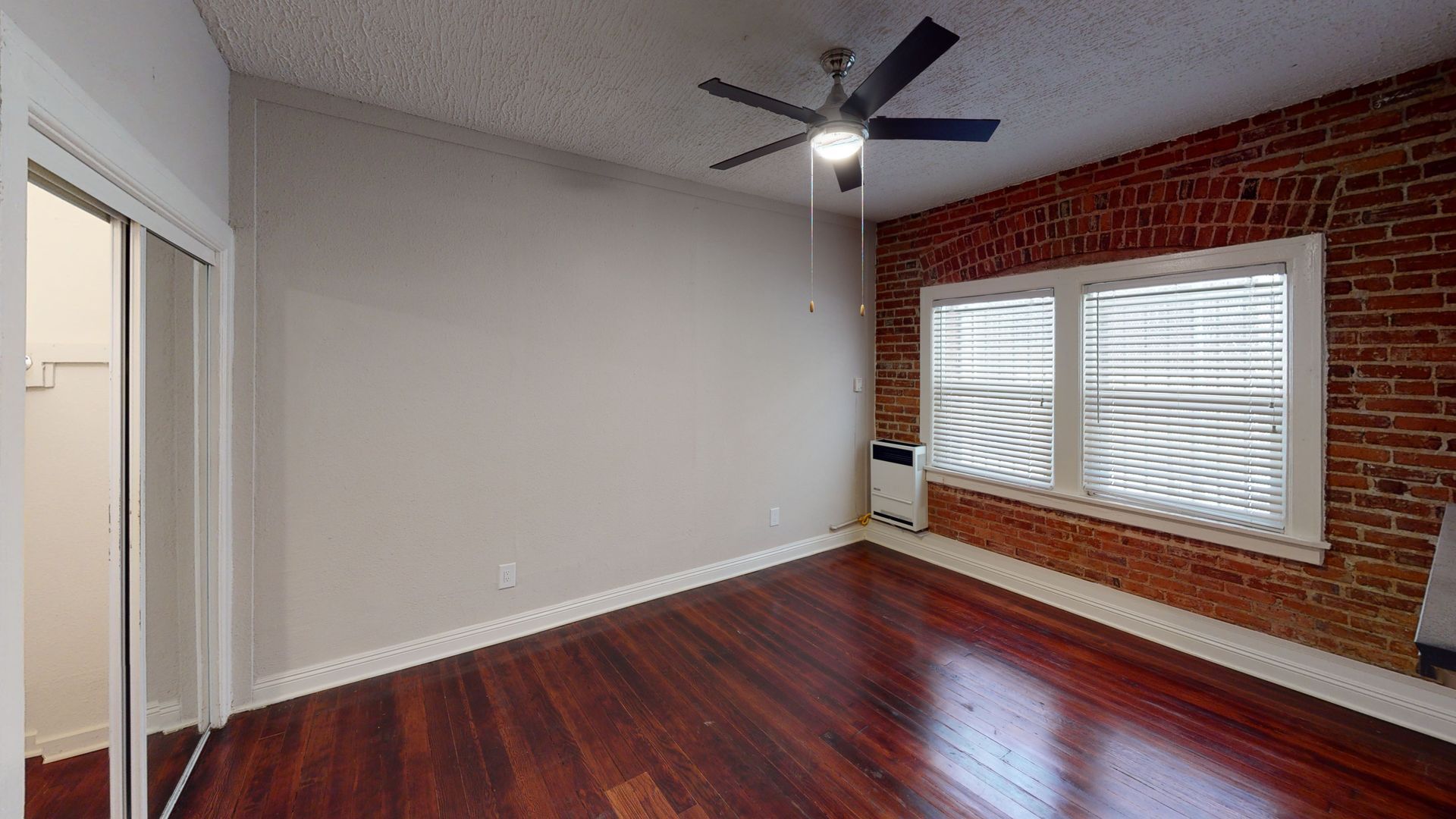Empty room with hardwood floors, exposed brick wall with window, and closet. Ceiling fan.