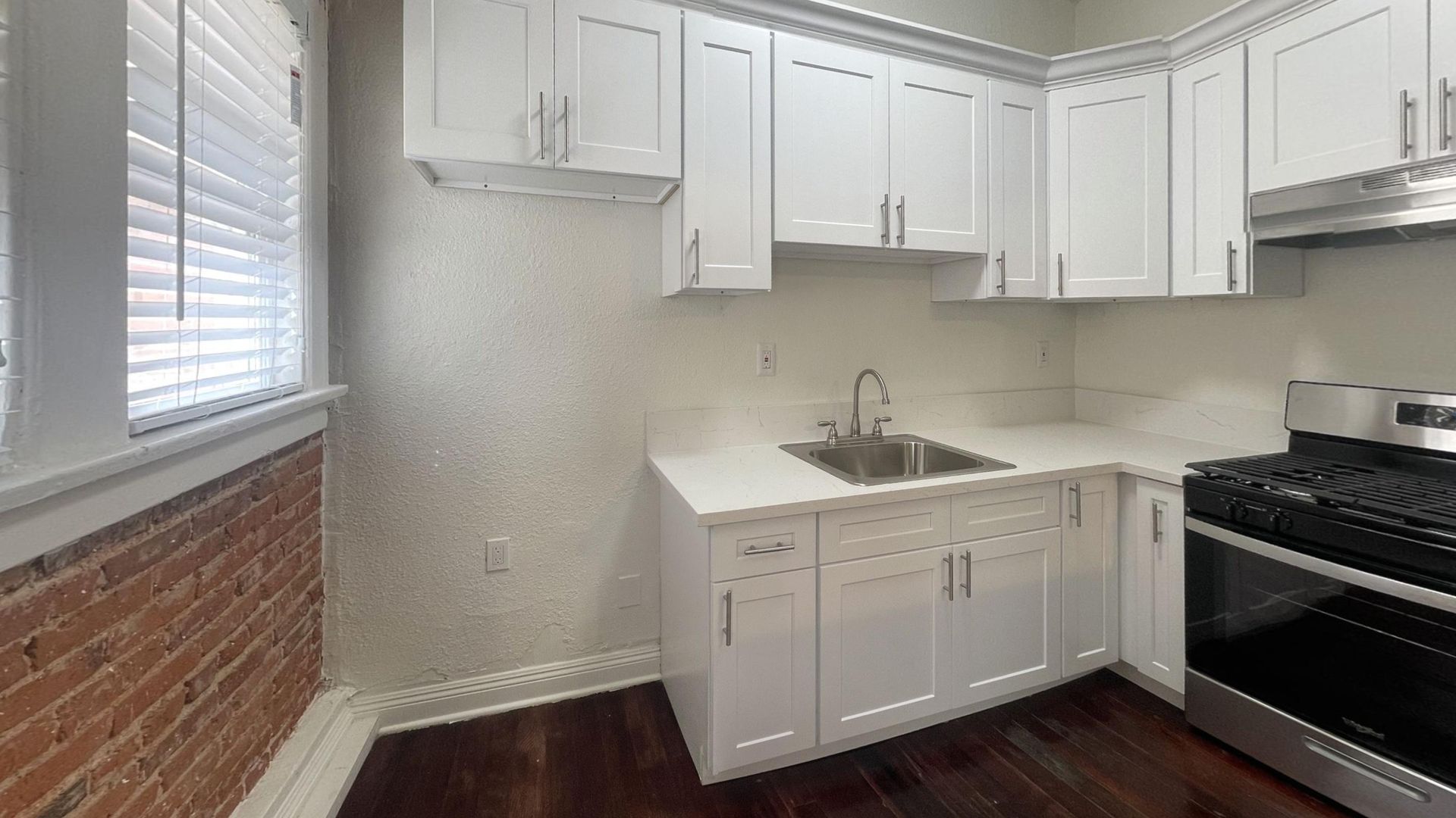 White kitchen cabinets, sink, stove, and window with blinds. Exposed brick wall on the left.