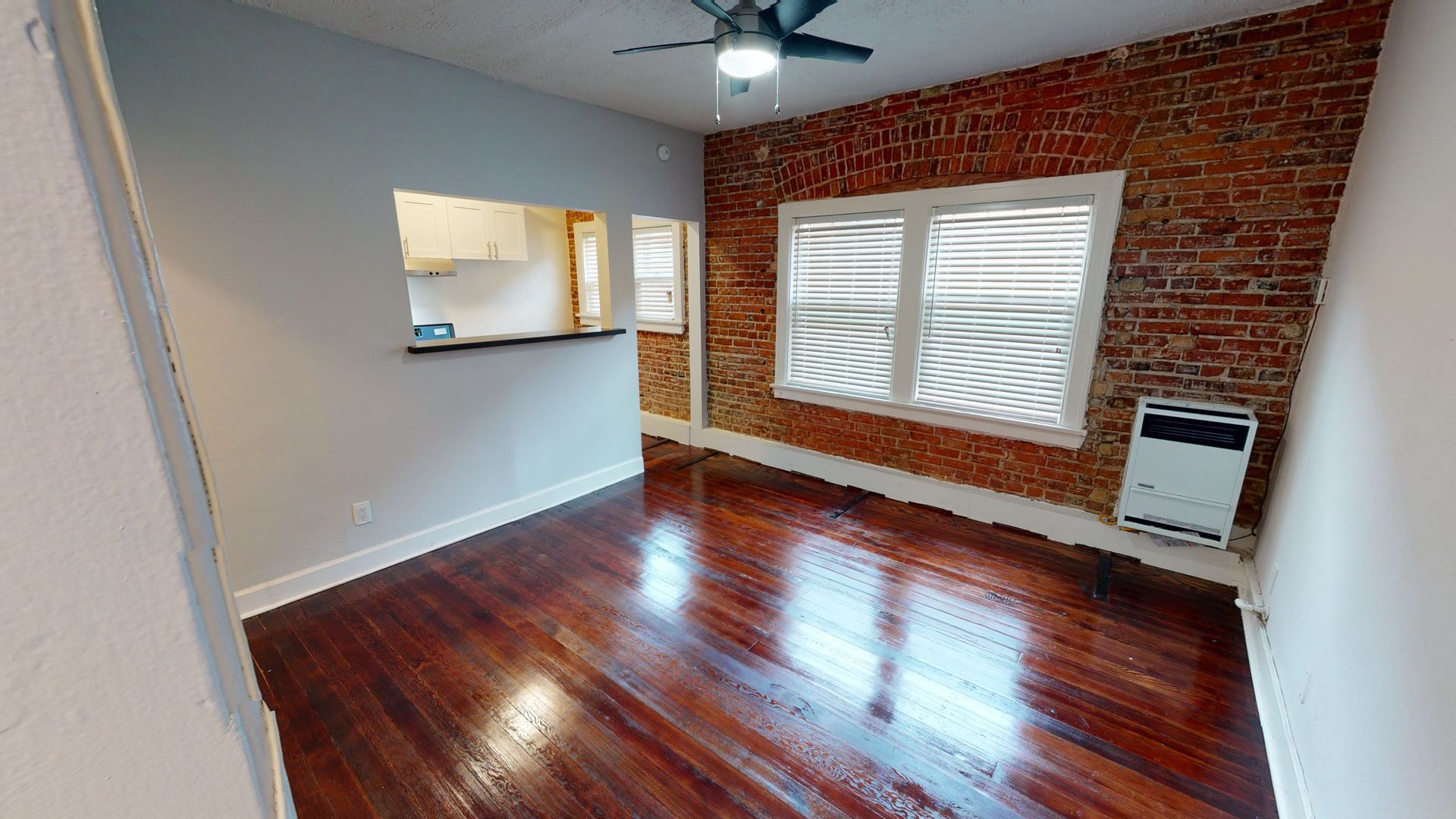 Interior view of a room with exposed brick wall, window, hardwood floors, and a small opening to a kitchen area.