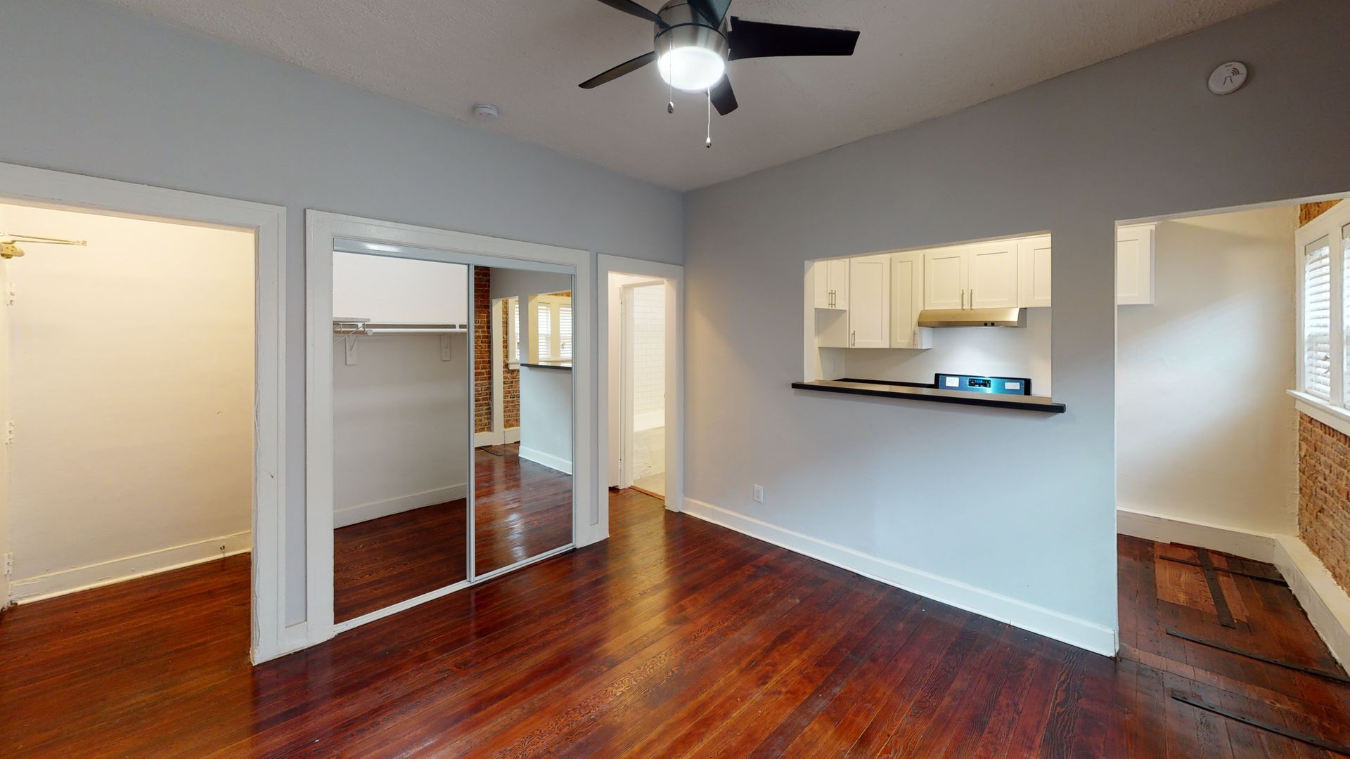 Empty apartment interior with hardwood floors, kitchen pass-through, and ceiling fan.