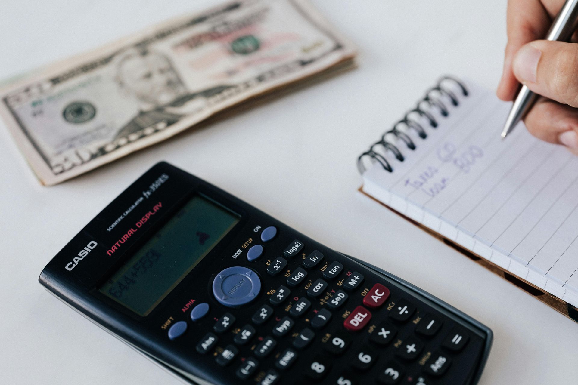 Calculator, cash, notebook, and hand writing on a white surface.