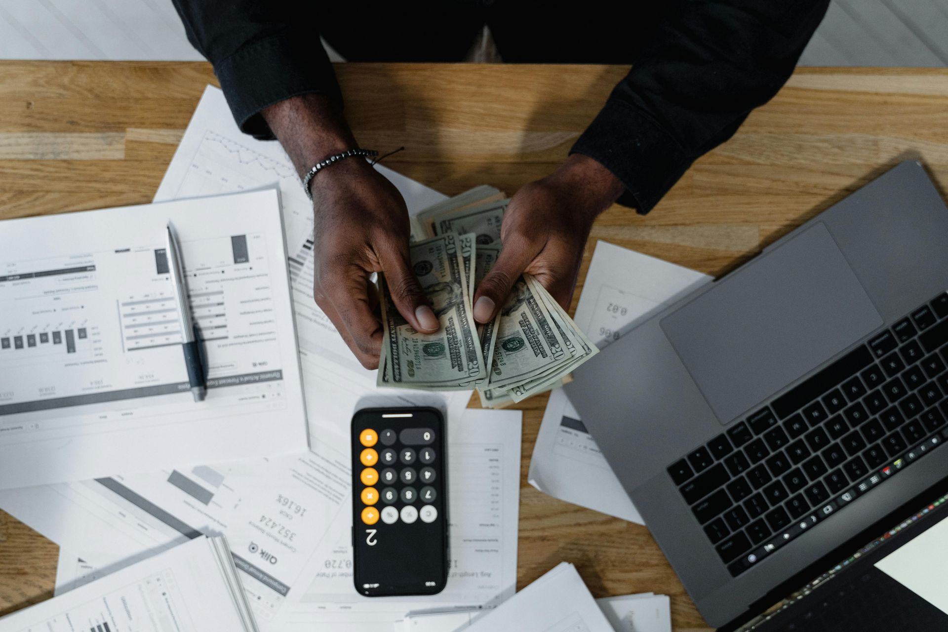 Hands counting cash over documents, calculator, and laptop on a wooden desk.