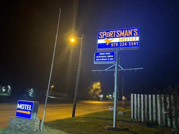 Sign for Sportsman's Motel at night, lit by streetlights. Includes phone number, WiFi, and directional sign.
