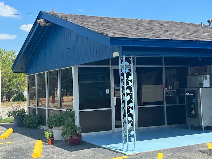 Blue building with large windows and a gray roof, in a parking lot on a sunny day.