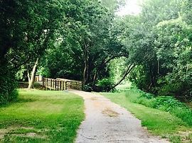 Path through a green park leads to a wooden bridge over a stream.