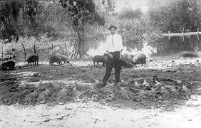 Man stands with a rifle before dead animals and pigs in a field near a body of water.