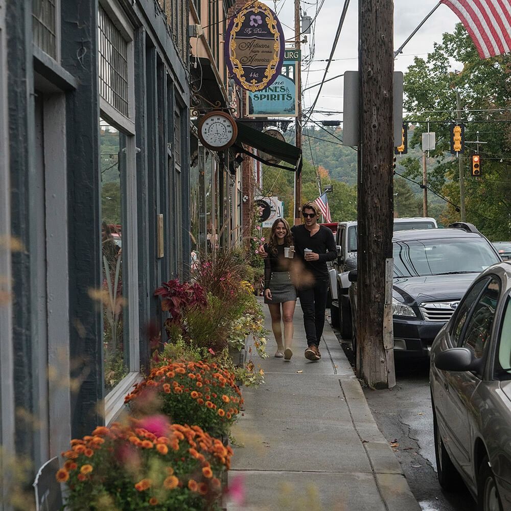 Two people walk down a sidewalk lined with flower pots, cars parked on the street. A shop with a sign and clock is visible.