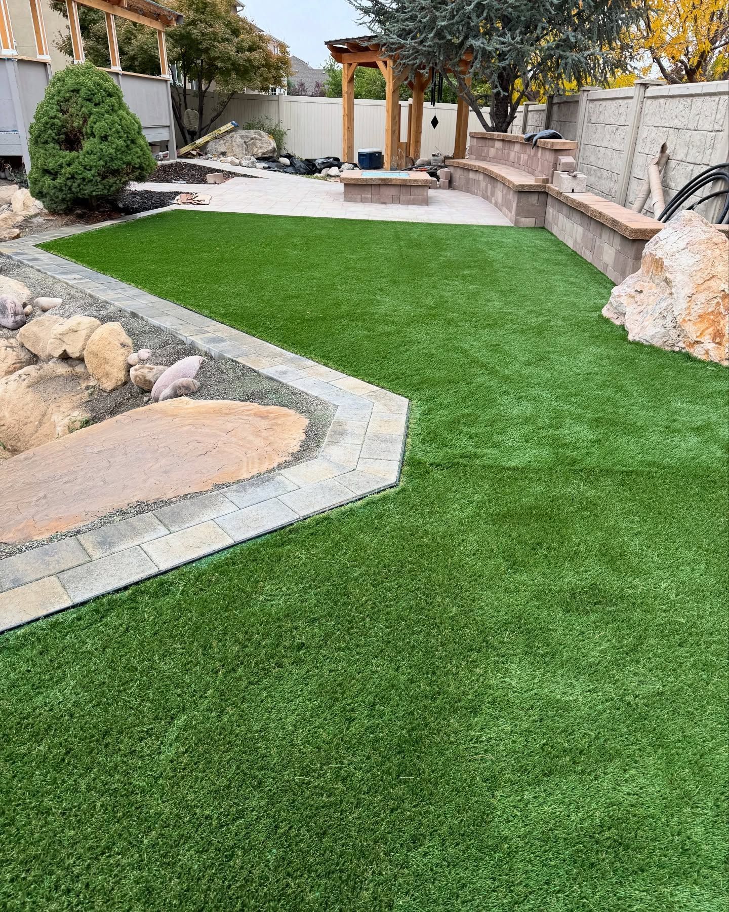 Green artificial turf in a backyard with a stone border, seating, and a fire pit. A rock and foliage border the yard.