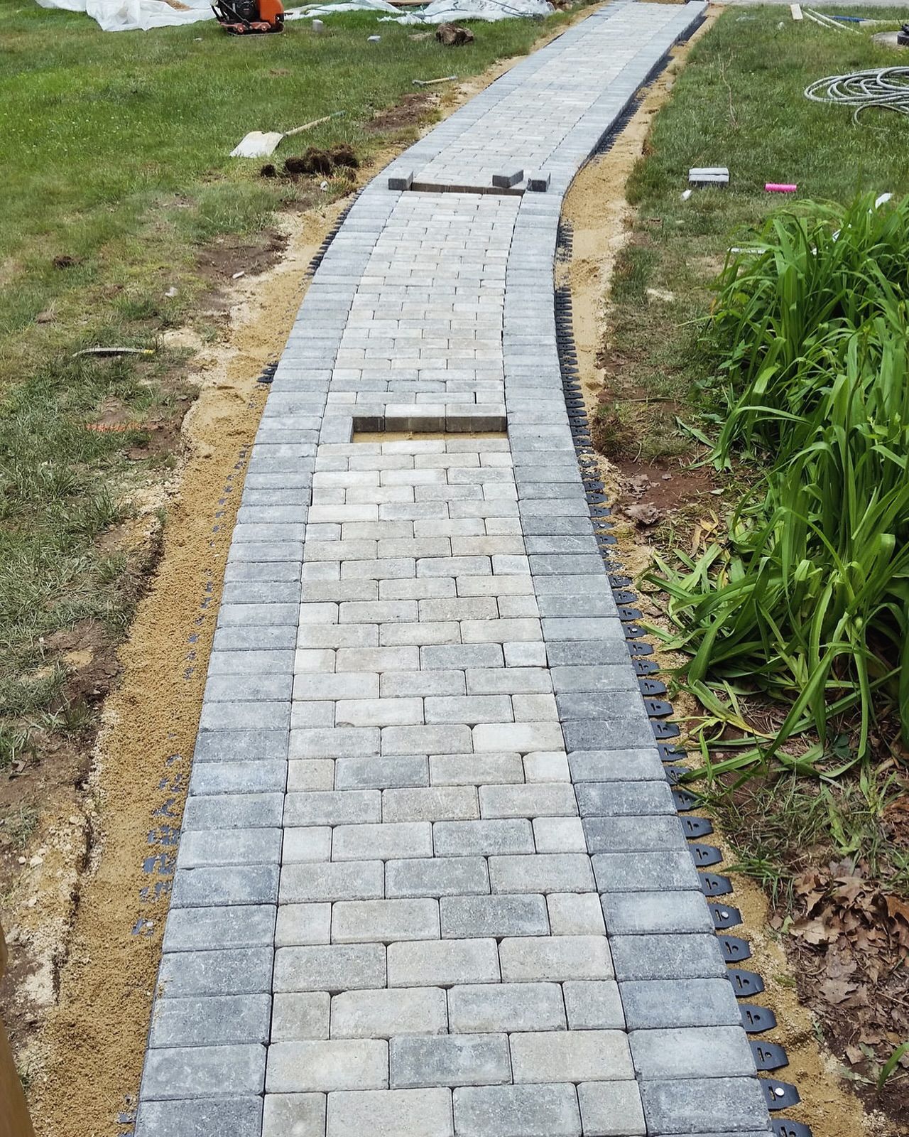 Brick pathway under construction, bordered by darker gray bricks and tan sand. Green grass surrounds the path.