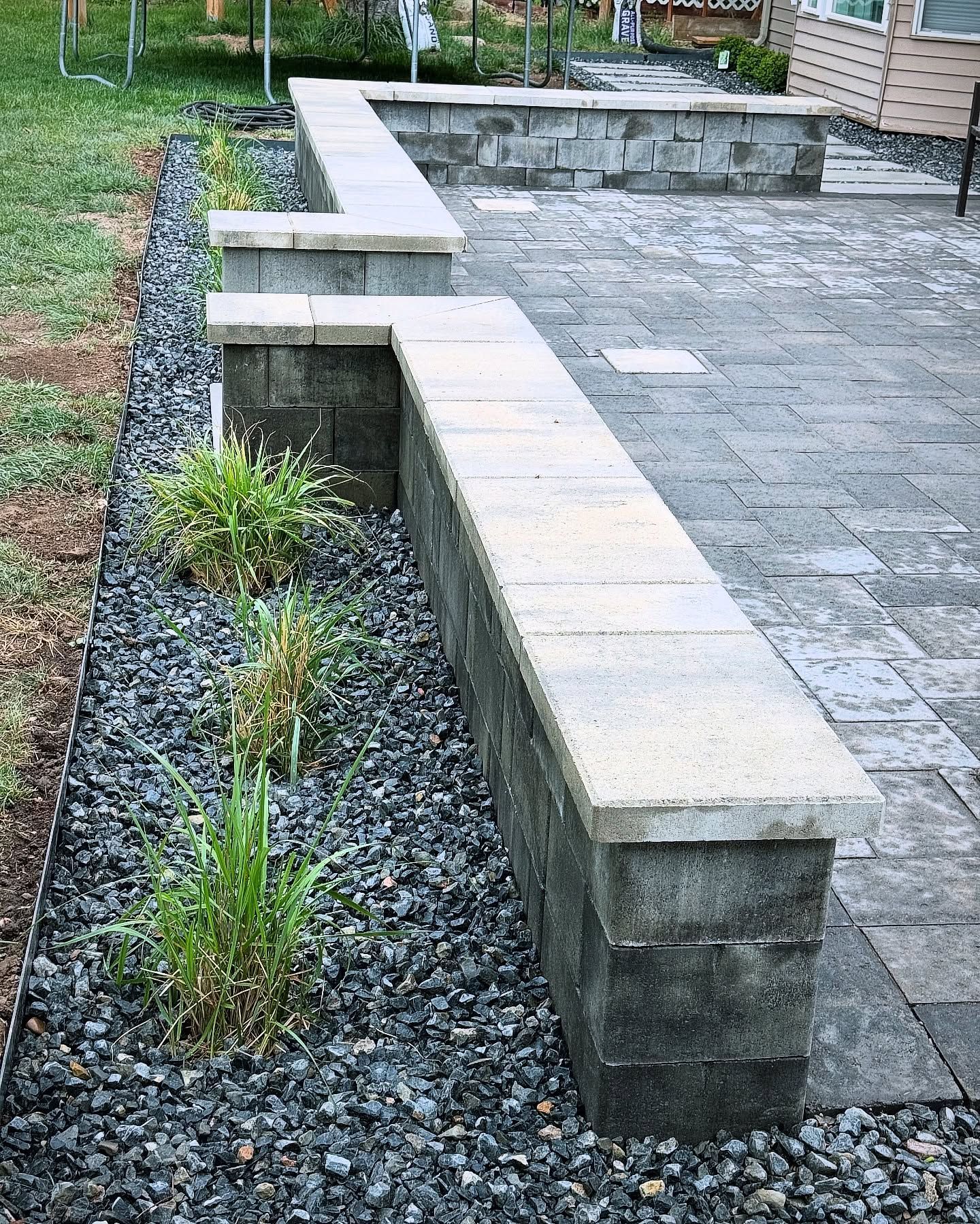 Long, low concrete retaining walls with pale caps separate a patio from a rock-filled garden bed with sparse green plants.