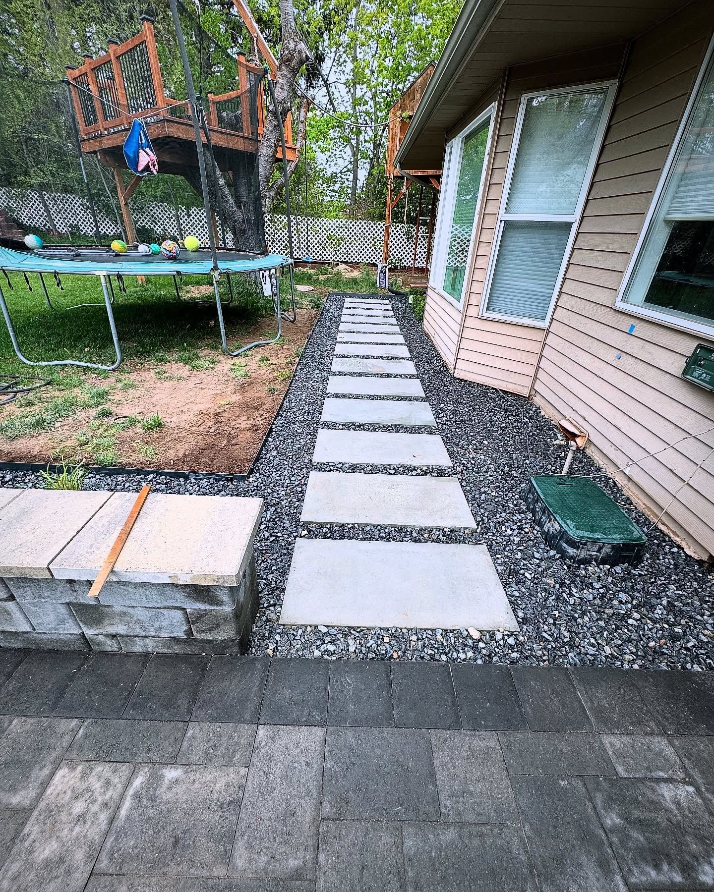 Stone walkway with gray pavers and black gravel alongside a light beige house with windows, leading to a treehouse in a backyard.