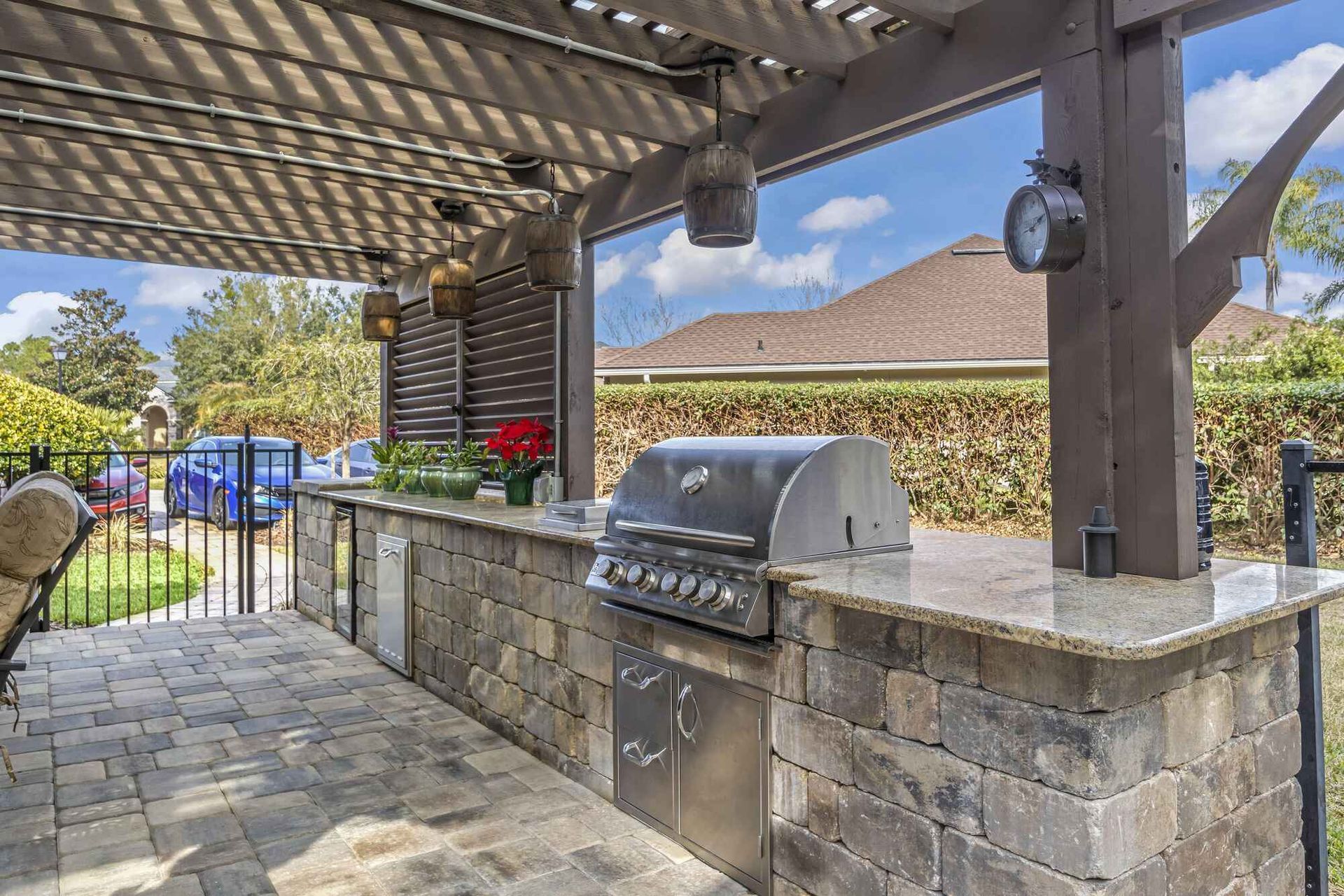Outdoor kitchen with a grill, stone facade, and a wooden pergola. The setting is a sunny patio with a view of houses and trees.