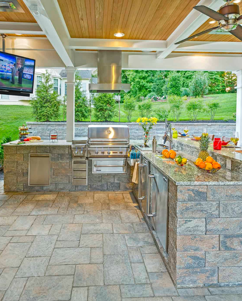 Outdoor kitchen with a grill, stainless steel appliances, and a granite countertop. The space includes a TV, and a brick-like floor.