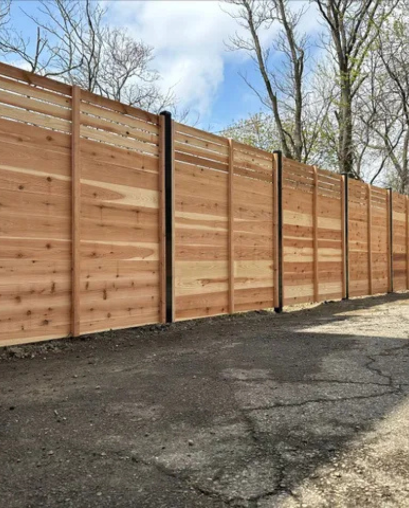 Wooden privacy fence with black posts lining a paved area, trees and a blue sky in the background.