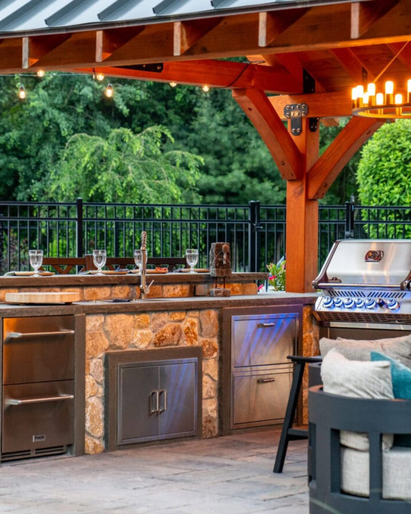 Outdoor kitchen with stone facade, stainless steel appliances, and pergola, overlooking a backyard with greenery.