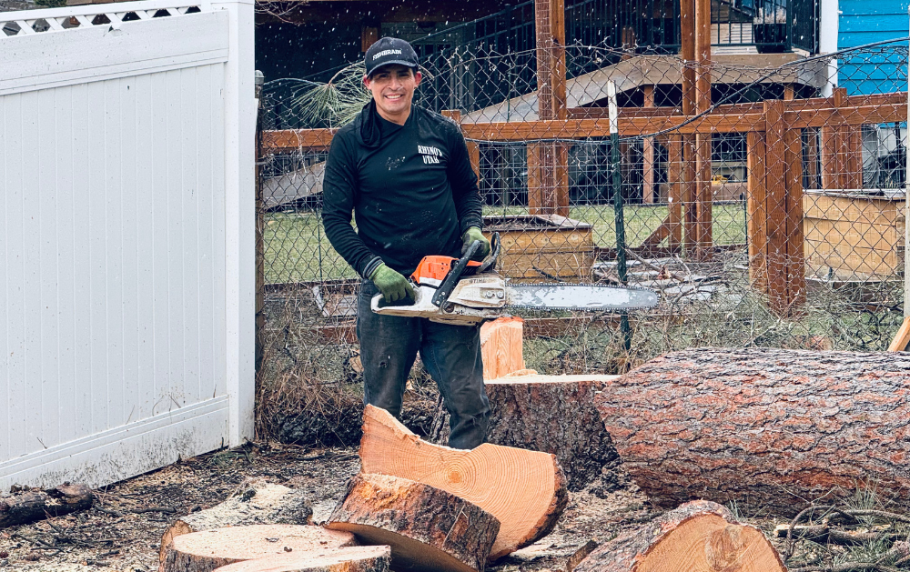 Man in black shirt and cap stands with chainsaw, cutting logs in a yard with a white fence.