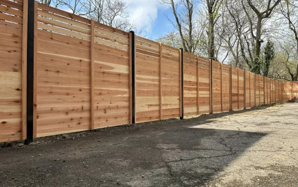 A long, horizontal wooden fence with dark posts in an outdoor setting. The ground is gravel and cracked pavement.