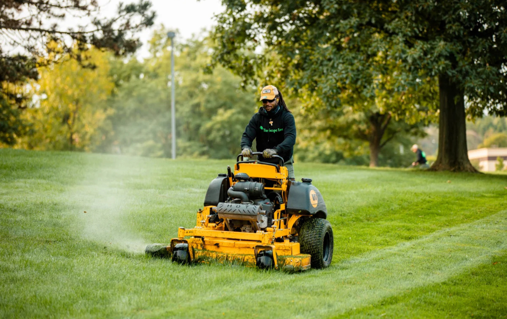 A person mowing a grassy hill with a yellow riding lawnmower on a sunny day. Trees and other landscaping are in the background.