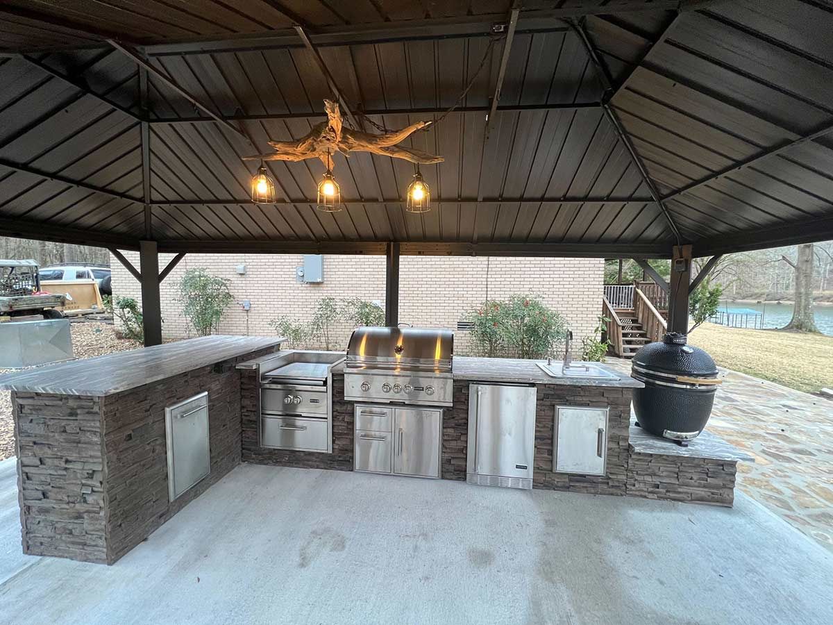 Outdoor kitchen under a brown metal gazebo. Features built-in grills, cabinets, sink, and a large ceramic smoker.
