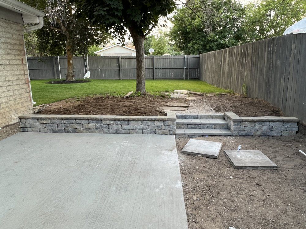 Backyard with a newly poured concrete patio and stone retaining wall steps leading up to a grassy area and a wooden fence.