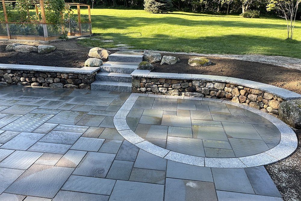 Stone patio with curved retaining wall, steps, and grassy area. Blue-grey paving stones contrast with the brown stone wall.